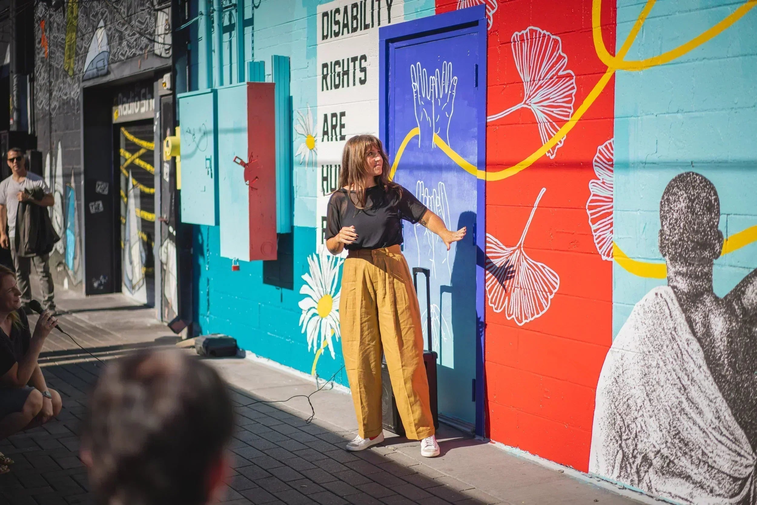Person wearing black t-shirt and mustard colored pants gestures to a bright colorful mural with the words "Disability Rights Are Human Rights"