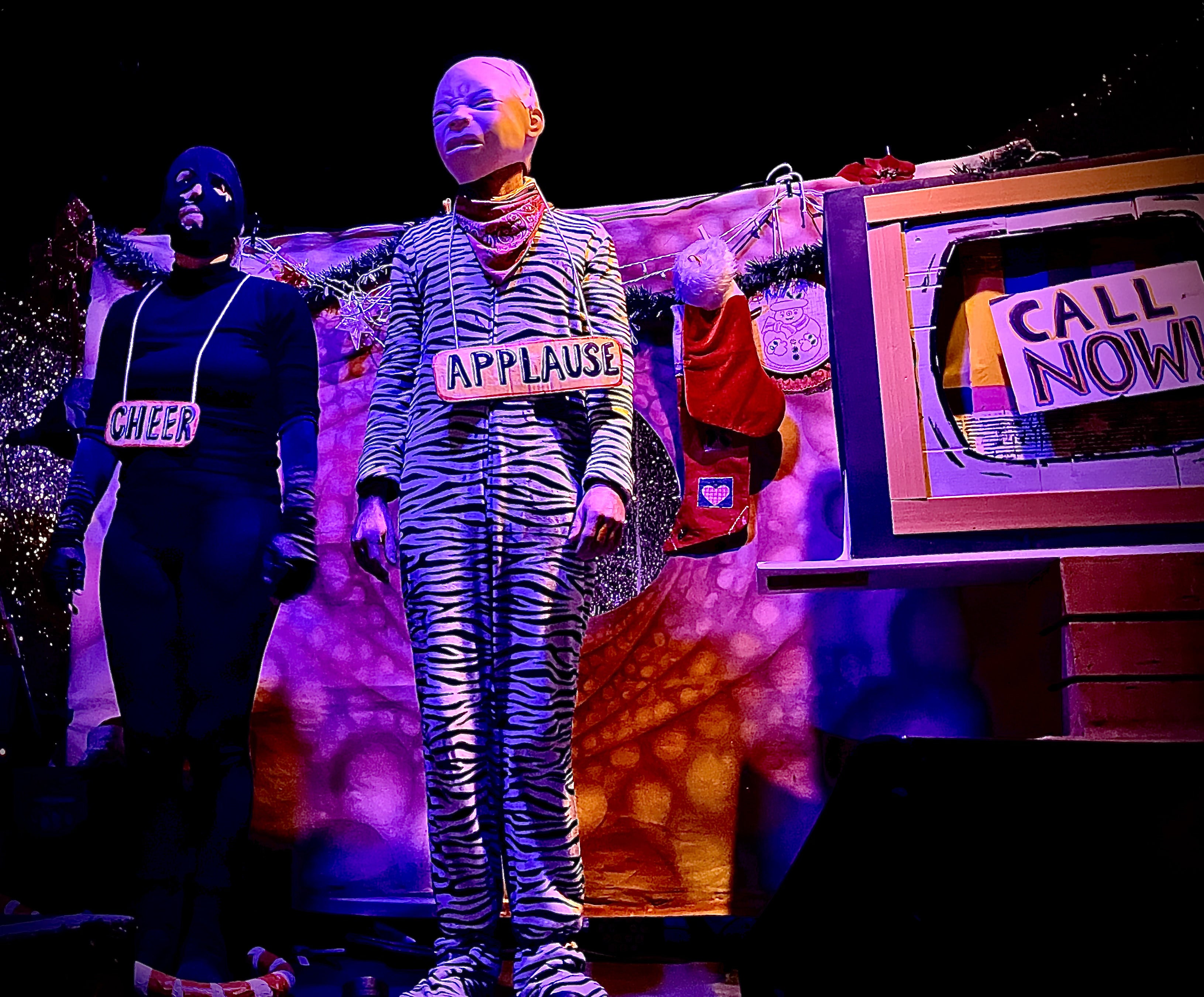 Person in zebra-striped costume with 'APPLAUSE' sign, standing in front of a colorful backdrop with text and props.