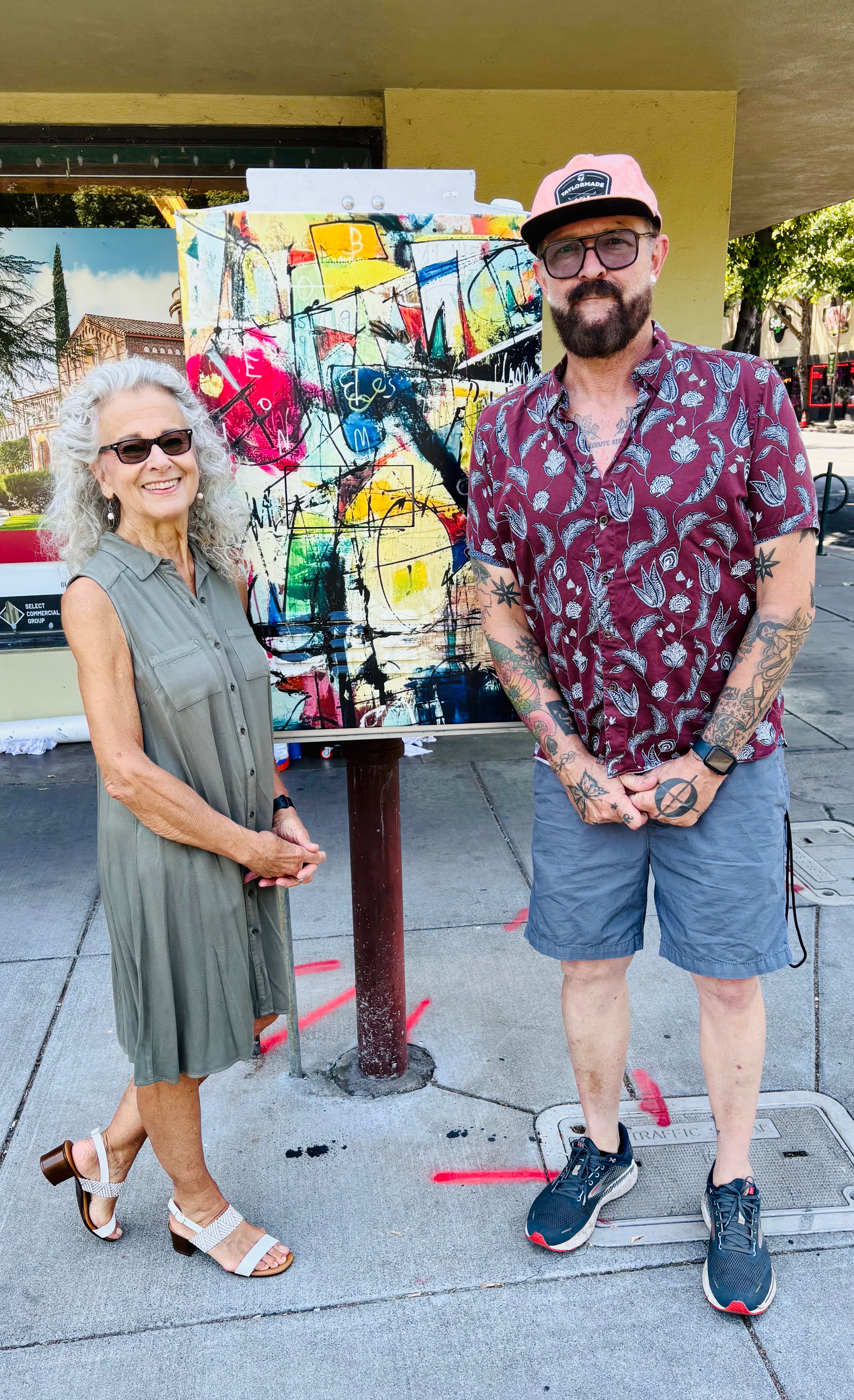 Two people standing next to a large painting outdoors.